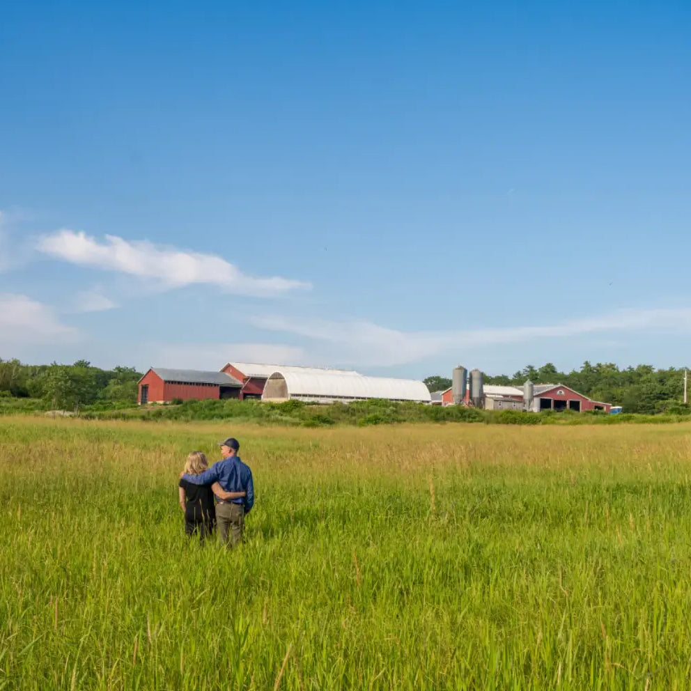Two people standing in a grassy field with a barn and trees in the background.